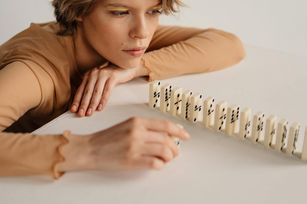 Dlaczego zawsze wszystko mi się sypie? Odkryj ukryty mechanizm Twoich porażek. A woman in focus, contemplating while playing with dominoes on a white table.