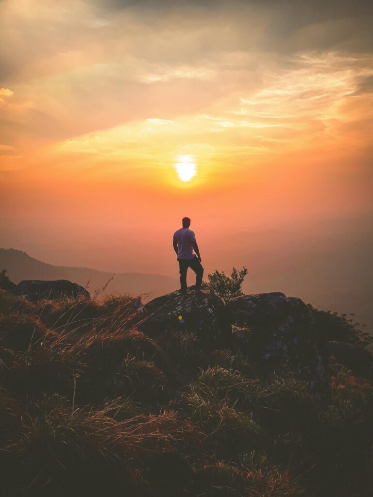 Moment, w którym zaczynasz wracać do siebie – odzyskaj swój wewnętrzny blask. A lone hiker admires a stunning sunset view from Ponmudi Peak, creating a serene landscape scene.